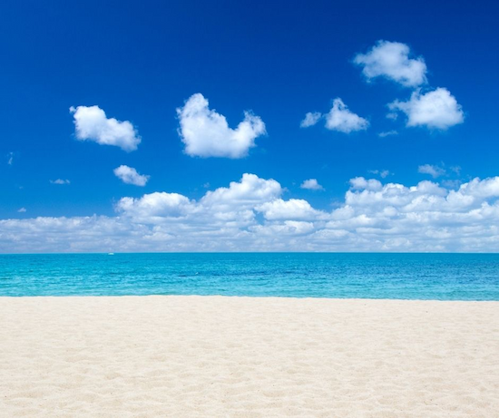 Heller Sandstrand vor türkisfarbenem Meer unter einem blauen Himmel mit weißen Wolken
