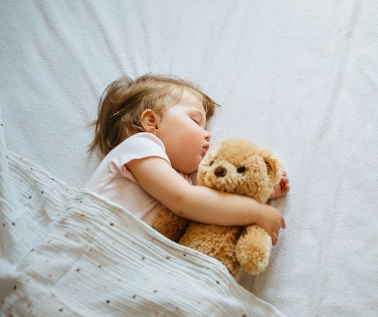 Ein kleines Kind schläft unter einer Decke und hält einen Teddybären im Arm.