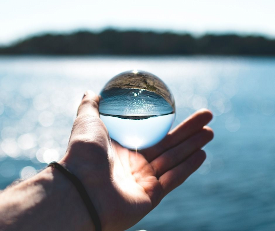 Eine Hand hält eine transparente Glaskugel vor einem Gewässer, in der die Umgebung spiegelverkehrt sichtbar ist.