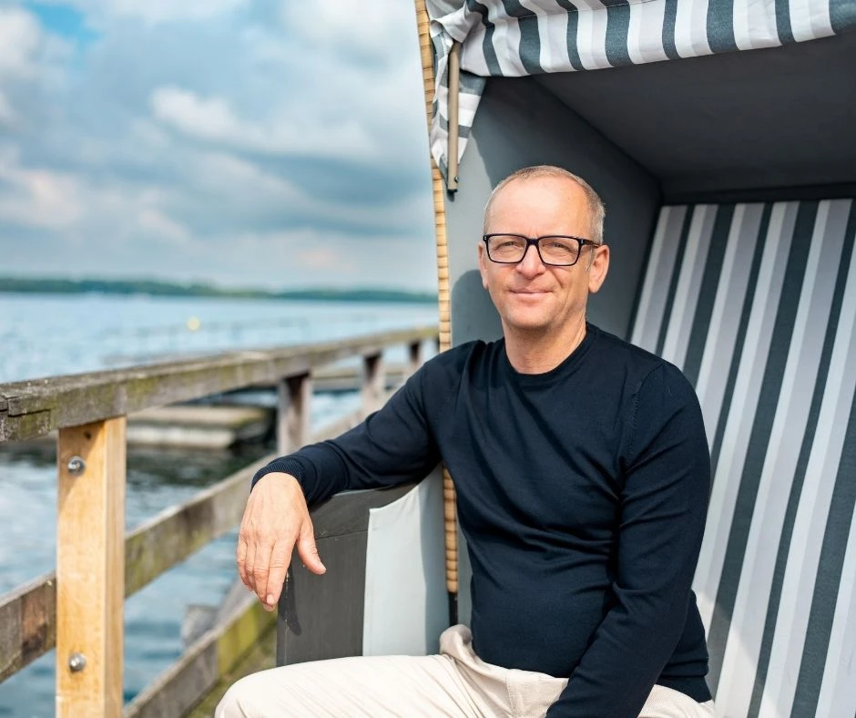 Eckart Julitz sitzt entspannt in einem Strandkorb am Meer und blickt ruhig in die Kamera.