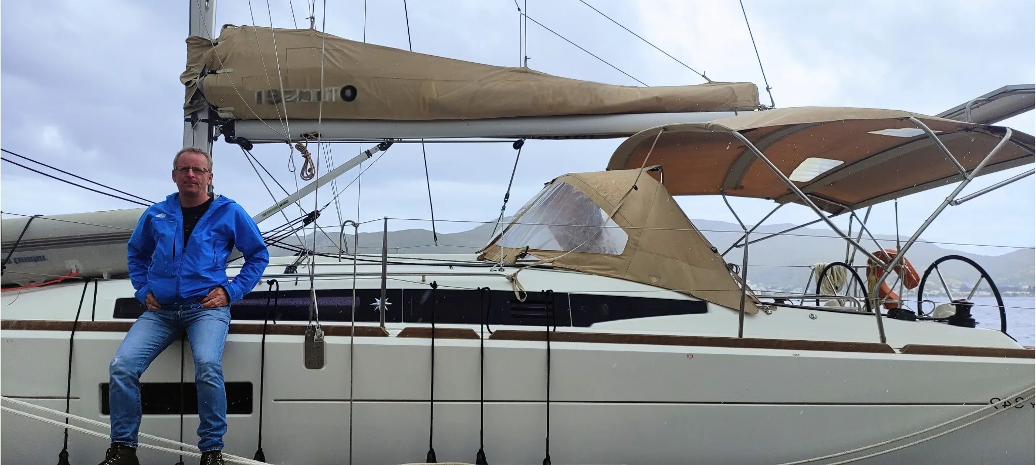 Eckart Julitz sitzt auf einem Segelboot vor Anker, im Hintergrund Mast, Segel und Küstenlandschaft